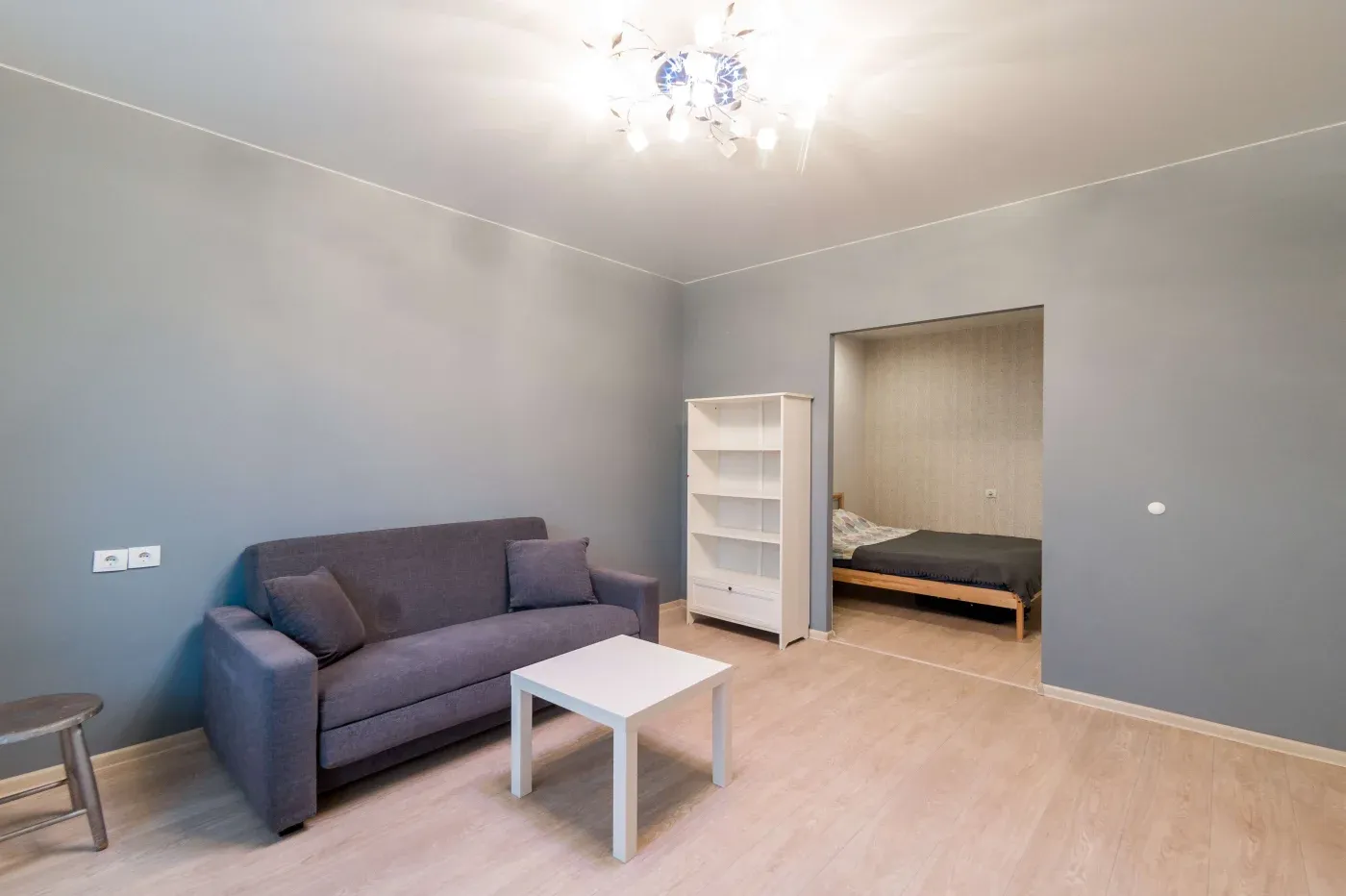 A minimalist living room with gray walls features a gray sofa, white coffee table, and bookshelf. A bright chandelier above. Bedroom view with a bed.