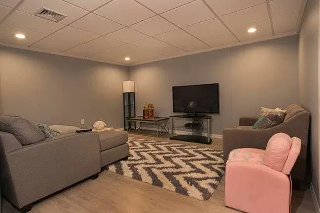 A cozy basement living room with muted gray walls and a white ceiling. It features a large gray sectional, a patterned rug, and a flat-screen TV on a stand. A pink child-sized chair and modern lamp add warmth to the space.
