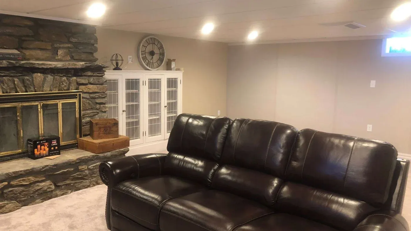 Spacious basement room with a leather sofa, stone fireplace, and glass cabinet. Warm lighting and neutral tones create a cozy, welcoming atmosphere.