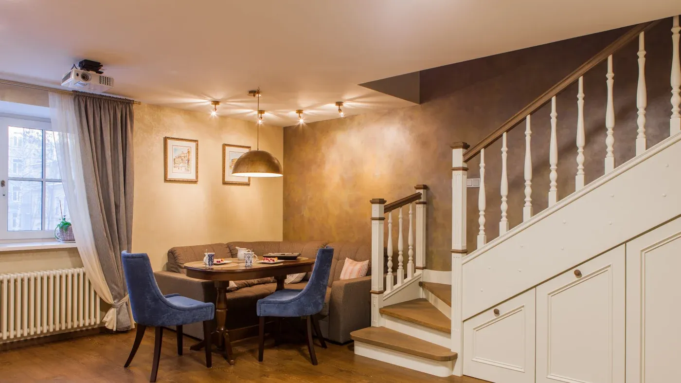 Cozy dining area with a wooden table, blue velvet chairs, and a beige corner couch. Warm lighting, artwork, stairs, and a window with drapes create a welcoming tone.