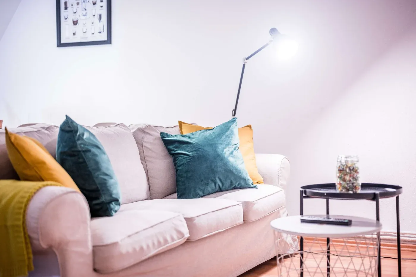 A cozy living room with a beige sofa adorned with teal and mustard pillows. A bright floor lamp illuminates a side table holding a glass jar under soft lighting.