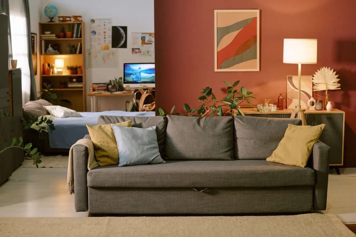 A cozy living room features a gray sofa with colorful pillows, a warm lamp, plants, and abstract art on a maroon wall, blending into a study area.