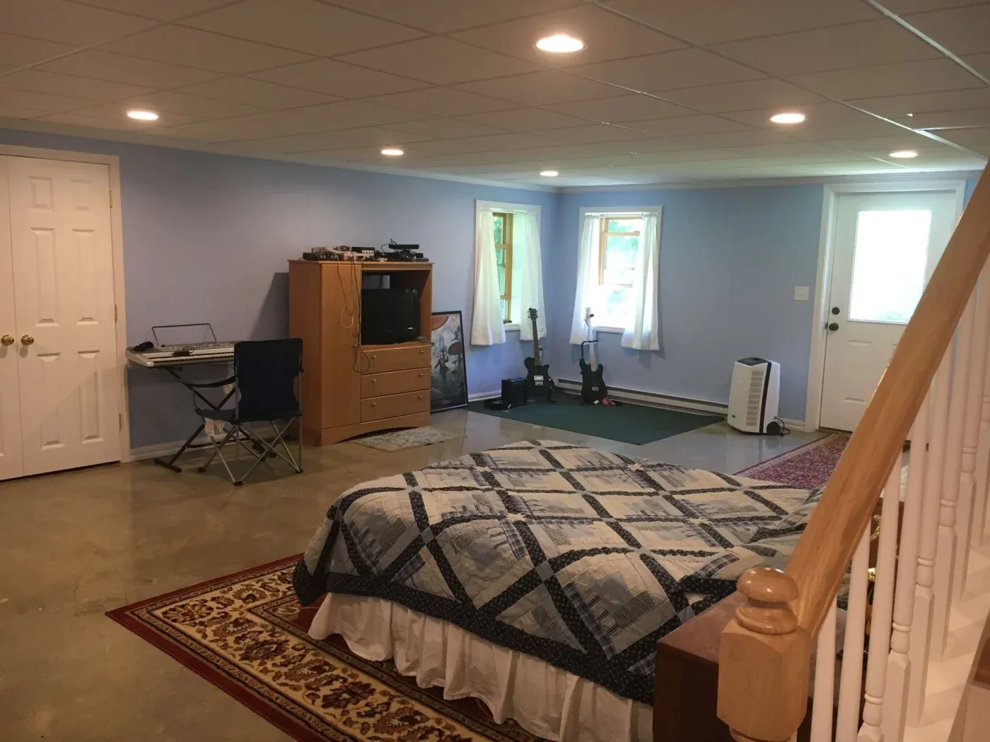 A cozy basement room with light blue walls features a bed with a blue and white quilt, a TV on a wooden dresser, two guitars by a window, and a folding chair at a small desk, creating a relaxed atmosphere.