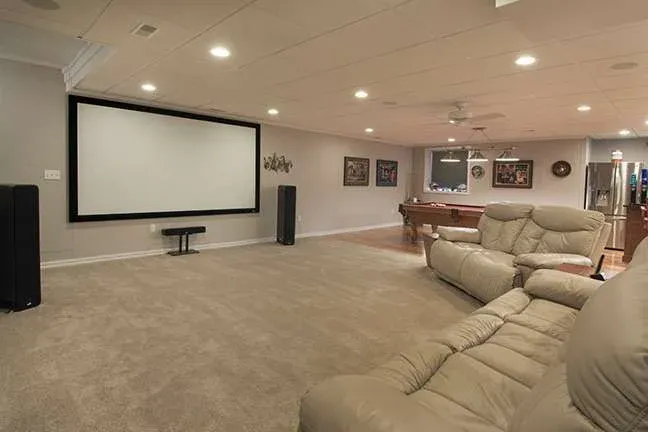 Spacious basement home theater with beige carpet and leather recliners. Large screen on one wall, pool table, and sports memorabilia create a cozy entertainment vibe.
