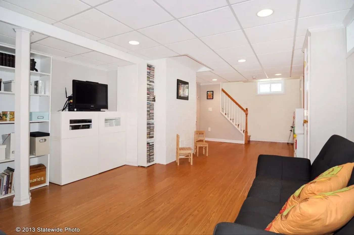 Spacious, tidy basement with wood flooring and white walls. Features a TV, bookshelves, a black sofa with colorful cushions, and stairs leading up. Calm, organized vibe.