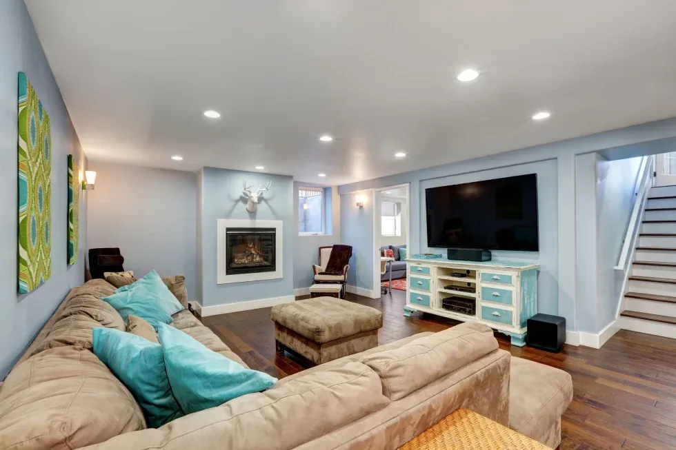 Spacious living room with light blue walls, tan sectional sofa, teal accents, and a wall-mounted TV. Features a modern fireplace and hardwood floors.