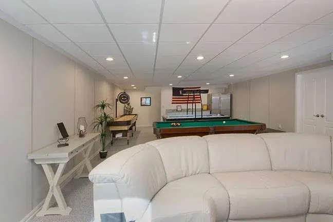 Basement game room with white sofa, pool table, and shuffleboard. Walls are light gray with soft lighting and an American flag on display.