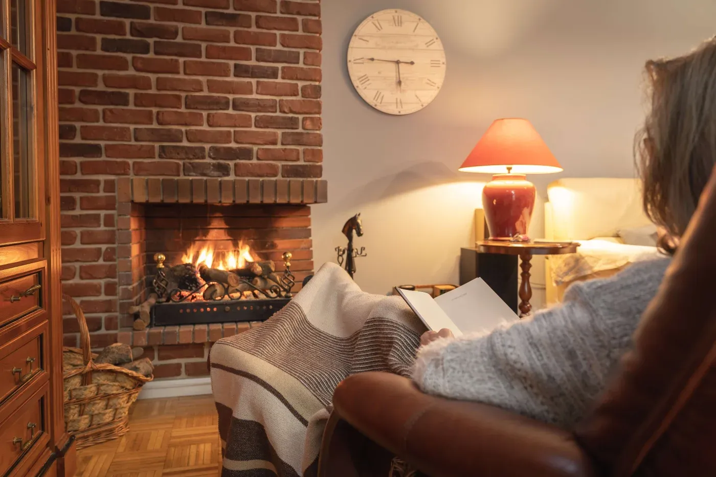A person relaxes in an armchair, wrapped in a blanket, reading by a lit brick fireplace. A warm lamp and clock adorn the cozy room.