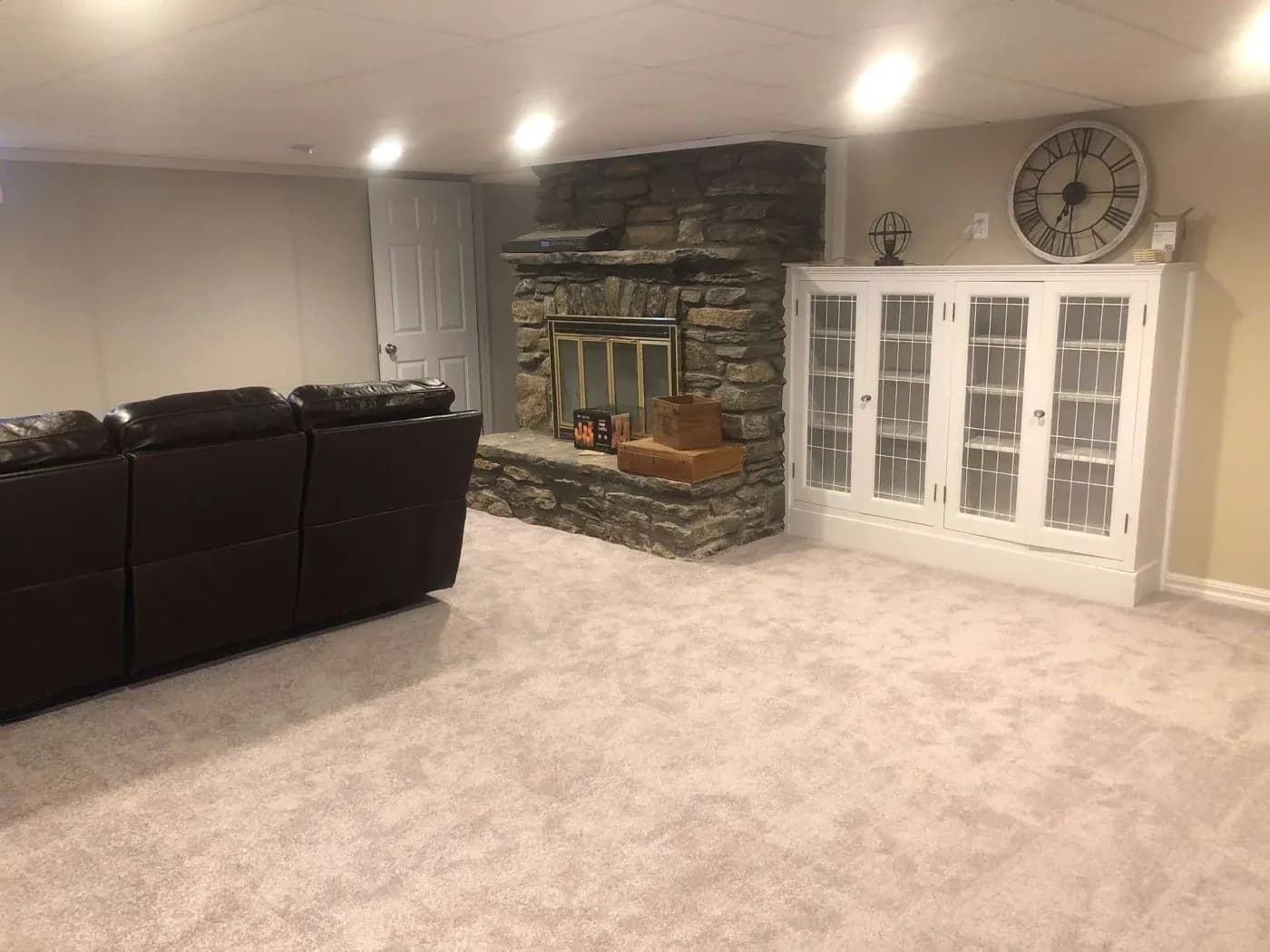 A cozy living room with plush carpet, featuring a brown leather sofa. A rustic stone fireplace with a brass screen is flanked by a white cabinet. A large clock decorates the wall, creating a warm and inviting ambiance.