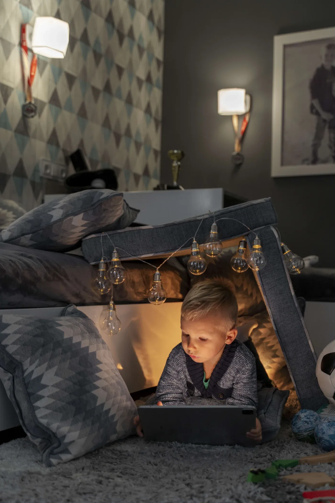 A young boy lies in a cozy pillow fort on a shaggy carpet, focused on a tablet. Warm string lights hang above, creating a calm, playful atmosphere.