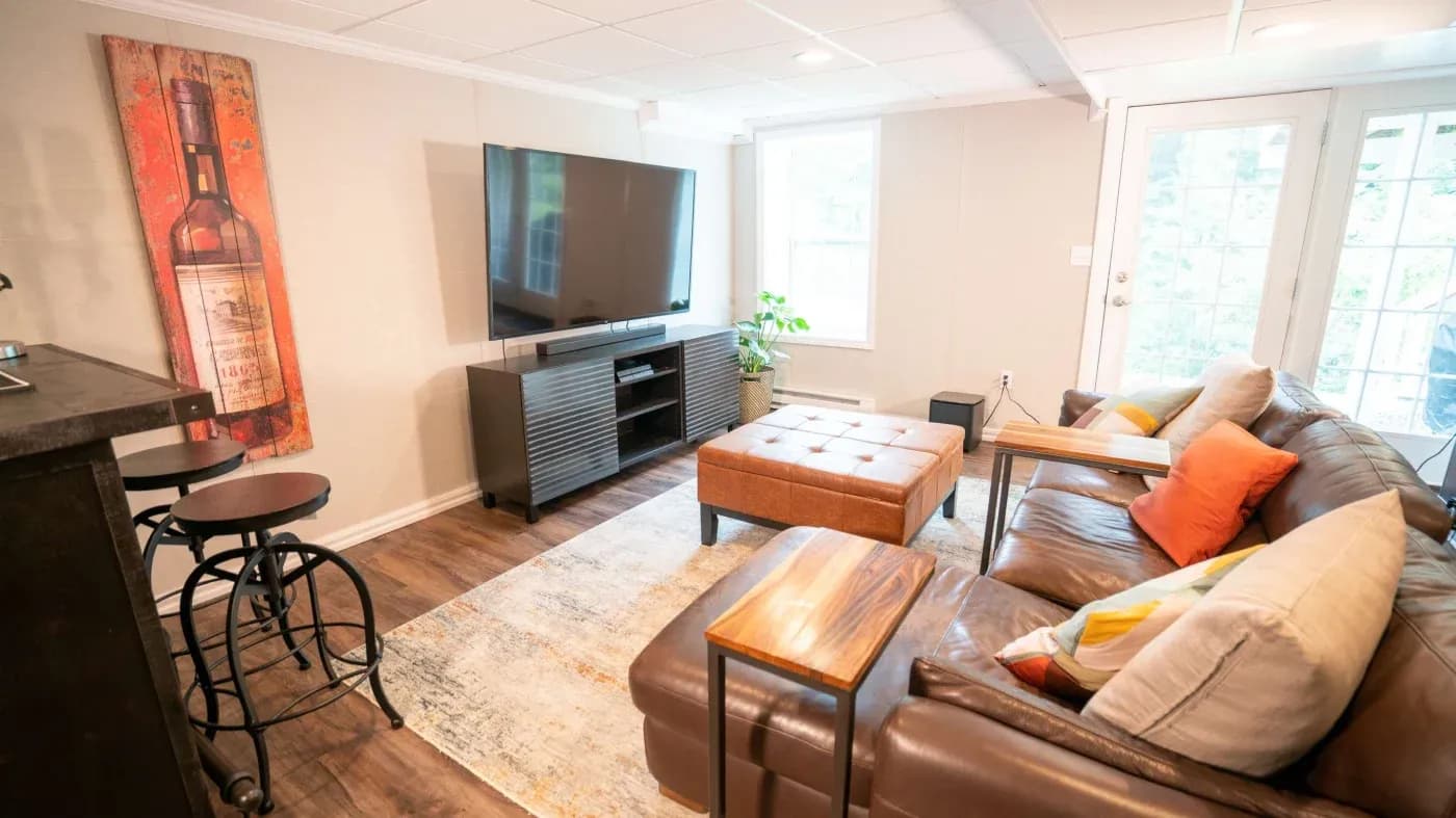 Cozy living room with a large brown leather sectional sofa, colorful cushions, a wooden ottoman, wall-mounted TV, and vintage wine bottle artwork.