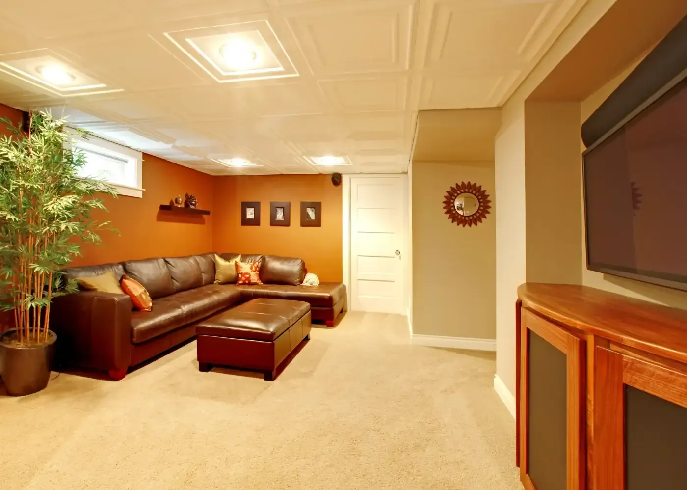 Cozy basement room with a brown leather sectional sofa and ottoman on beige carpet. Warm orange walls, ceiling lights, wall art, and a potted plant enhance comfort.