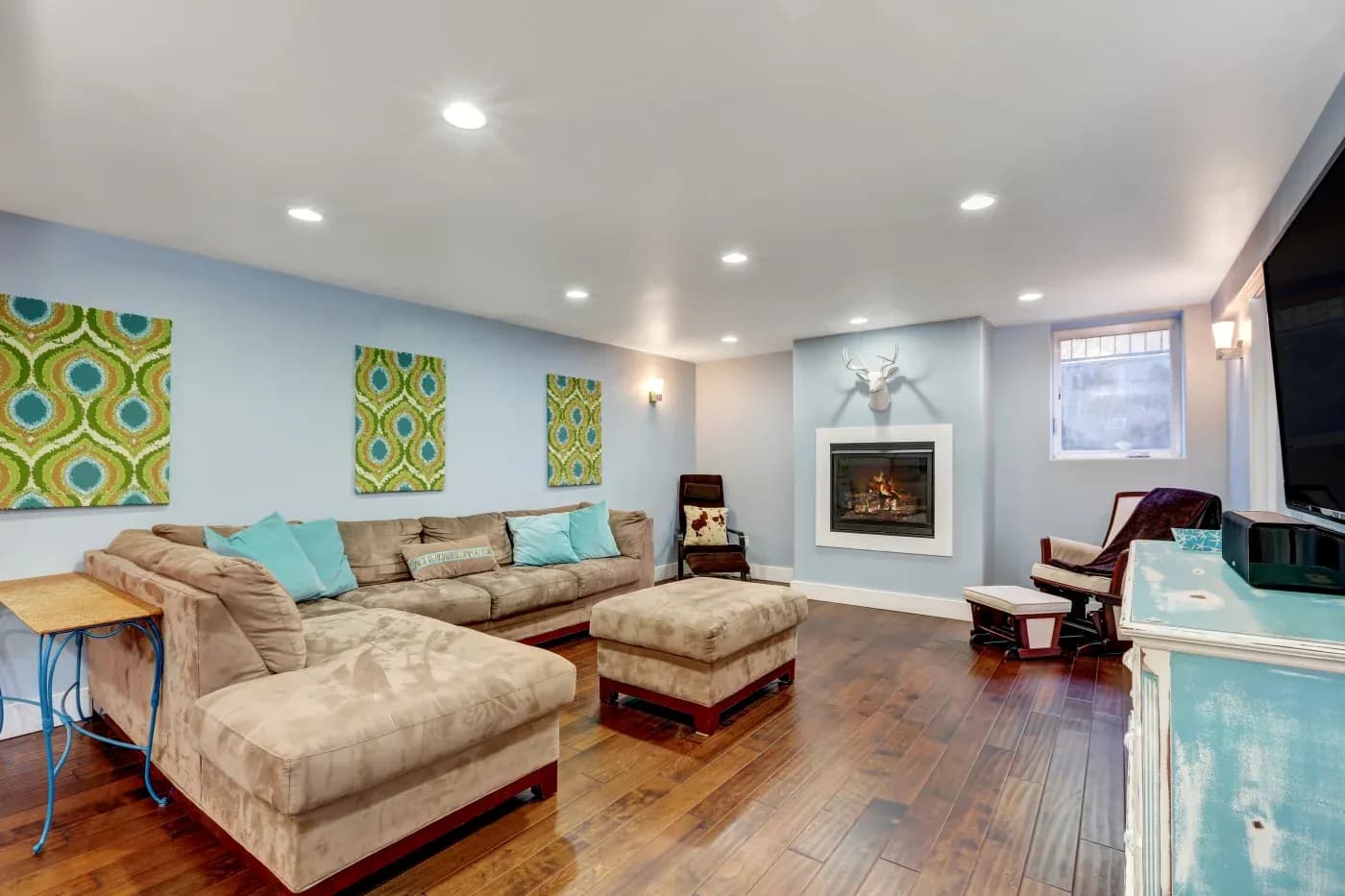 Modern living room with a tan sectional sofa, pastel blue cushions, patterned wall art, and a cozy fireplace. Warm lighting and wooden floors enhance comfort.