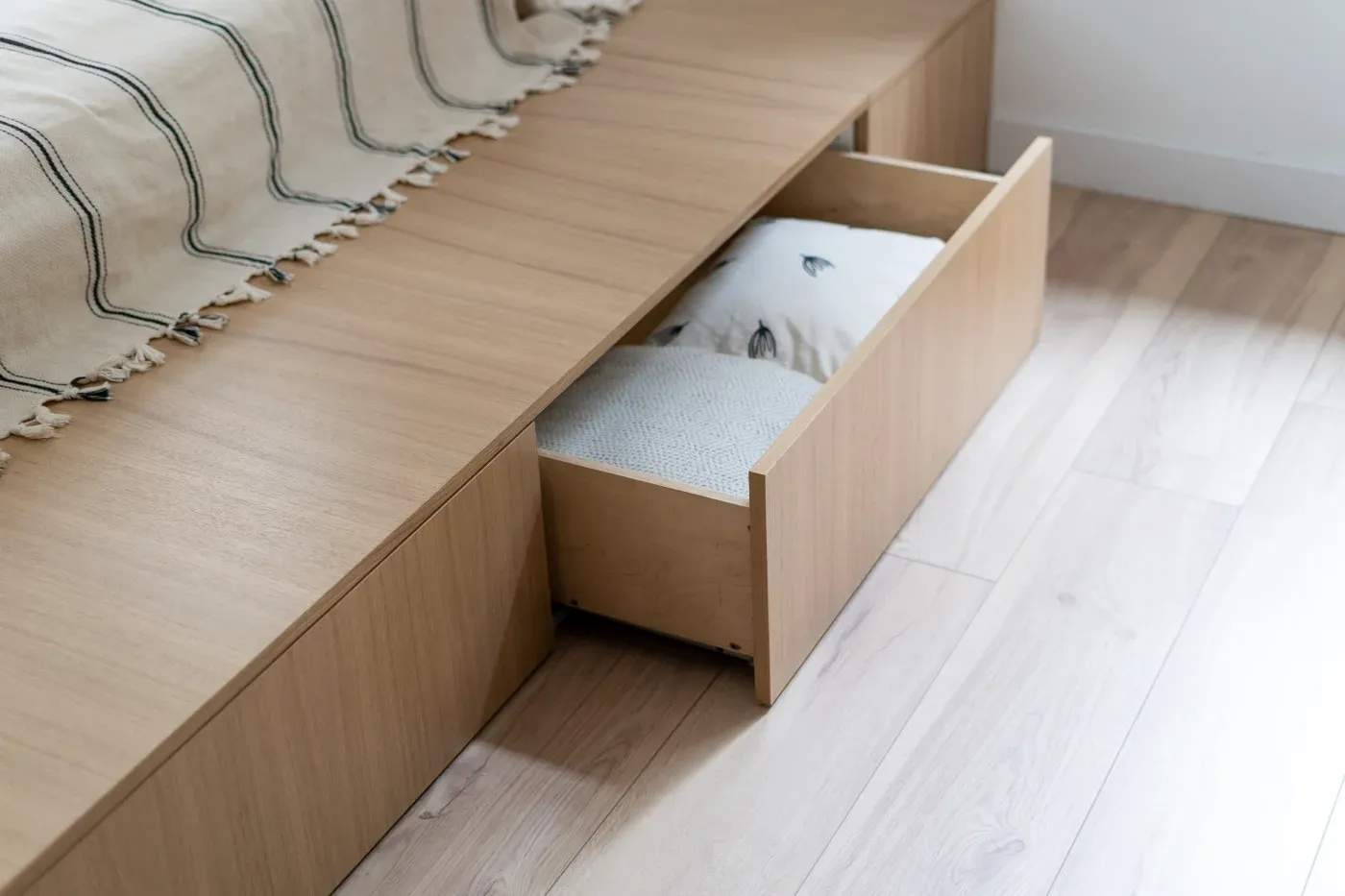 Wooden bed frame with an open drawer, revealing neatly folded blankets. A beige cover with green stripes drapes over the bed, creating a tidy, cozy ambiance.