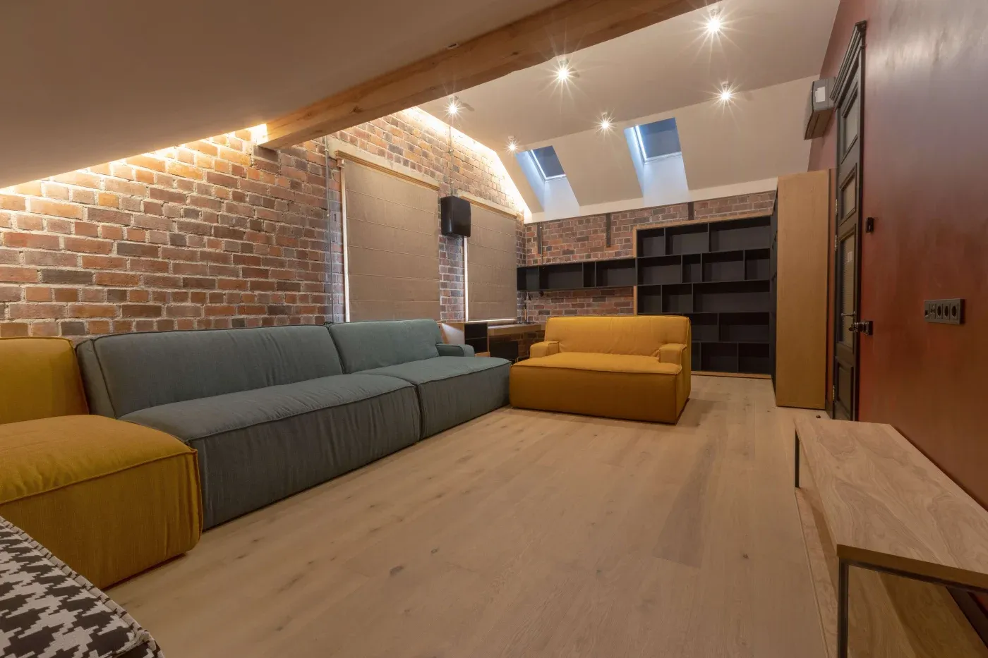 Spacious loft living room with exposed brick walls, wood floors, a blue and mustard sectional sofa, skylights, and built-in shelves under soft lighting.