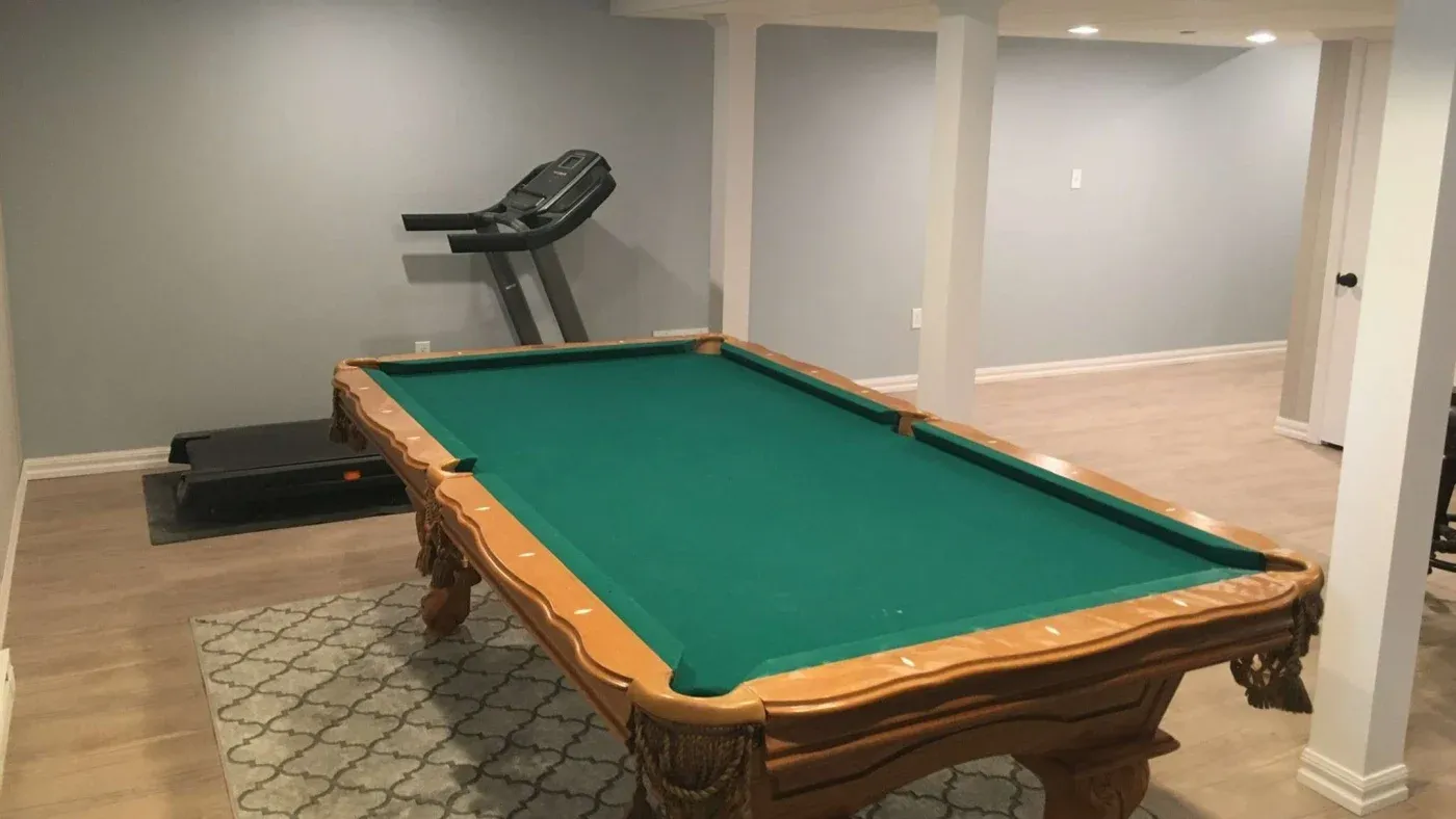 A spacious room with a green-felt pool table on a patterned rug. A treadmill is positioned against the gray wall in the background, creating a recreational vibe.