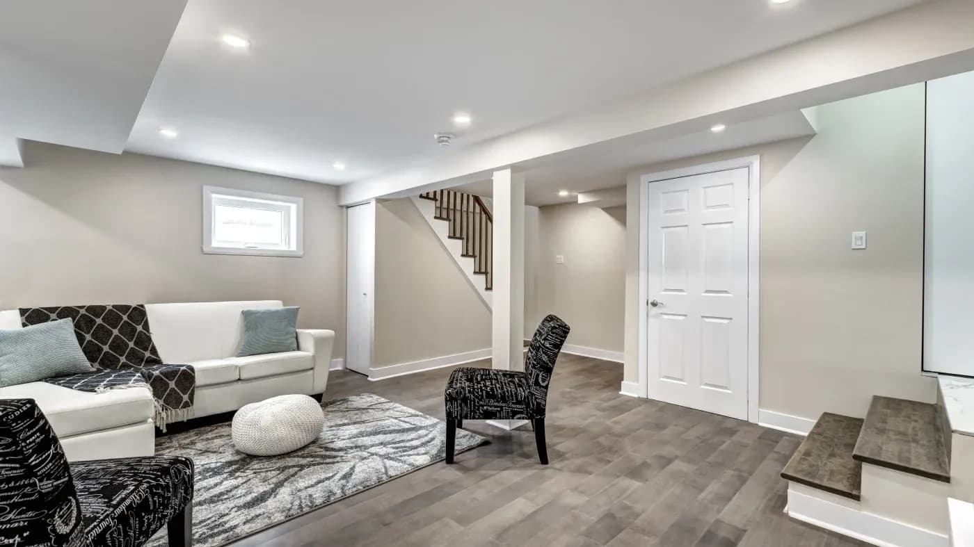 Modern basement living area with a white sofa, patterned armchairs, and a gray rug. A staircase and white door are visible, with soft recessed lighting.