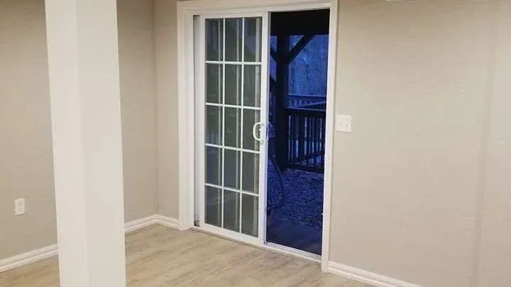 A room with beige walls and wood flooring features a glass sliding door slightly open, revealing a deck with a dimly lit outdoor scene at dusk.