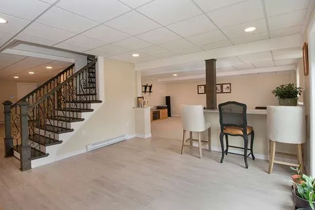 Bright basement room with neutral tones, modern decor, and a cozy vibe. Elegant stairs with iron railings, bar area with seating, and potted plants.