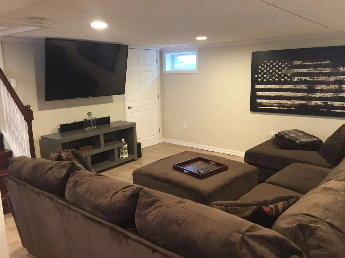 Cozy basement living room with a large sectional sofa and ottoman. A TV is mounted on the wall, and artwork of an American flag adds a patriotic touch.