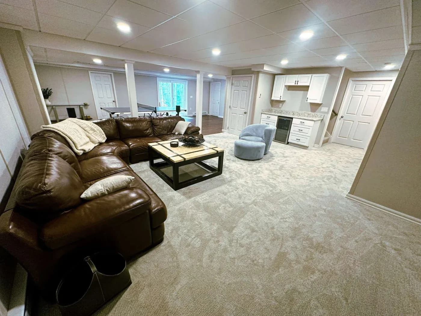 Spacious basement with plush carpet, featuring a large brown leather L-shaped sofa, cozy armchair, modern coffee table, and a kitchenette in the background.
