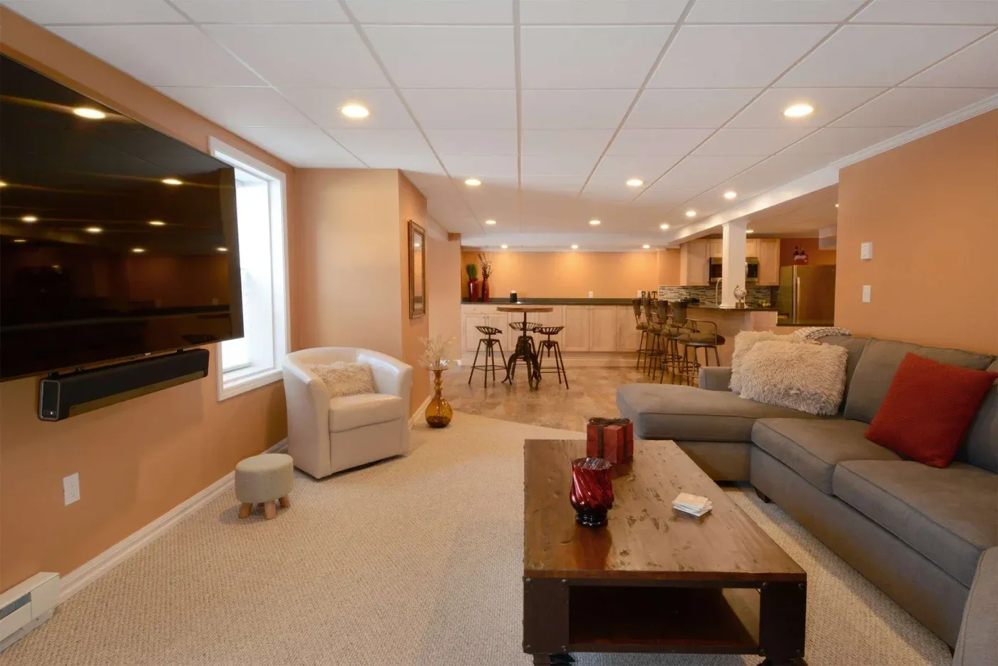 Spacious basement living area with warm lighting, a large gray sectional, flat-screen TV, cozy chair, and a bar with stools, creating a welcoming vibe.