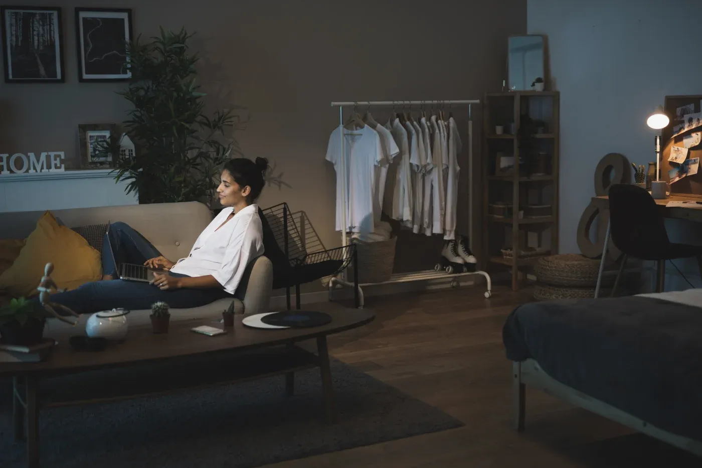 A woman relaxes on a gray sofa, holding a laptop in a cozy, dimly lit room. Nearby, a clothing rack with white shirts and a warmly lit desk create a serene, homey atmosphere.
