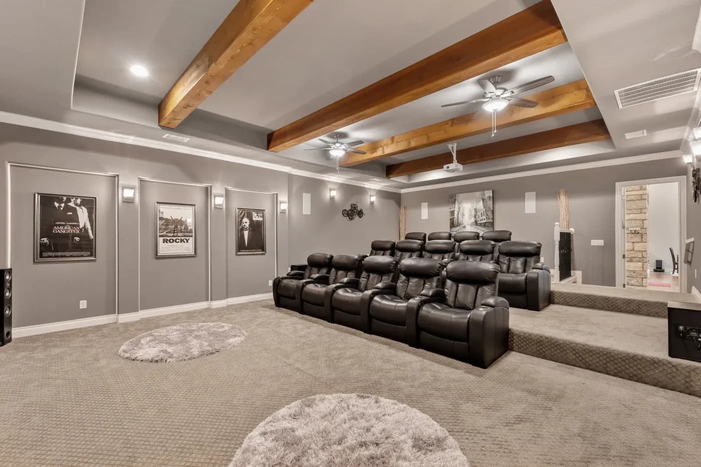 Luxurious home theater with tiered black leather recliners, framed movie posters on gray walls, wooden beams, ceiling fans, and a cozy carpeted floor.