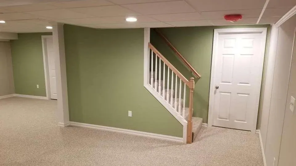 A basement room with light green walls, beige carpet, and white doors. A staircase with a wooden handrail leads upstairs, conveying a clean, empty space.