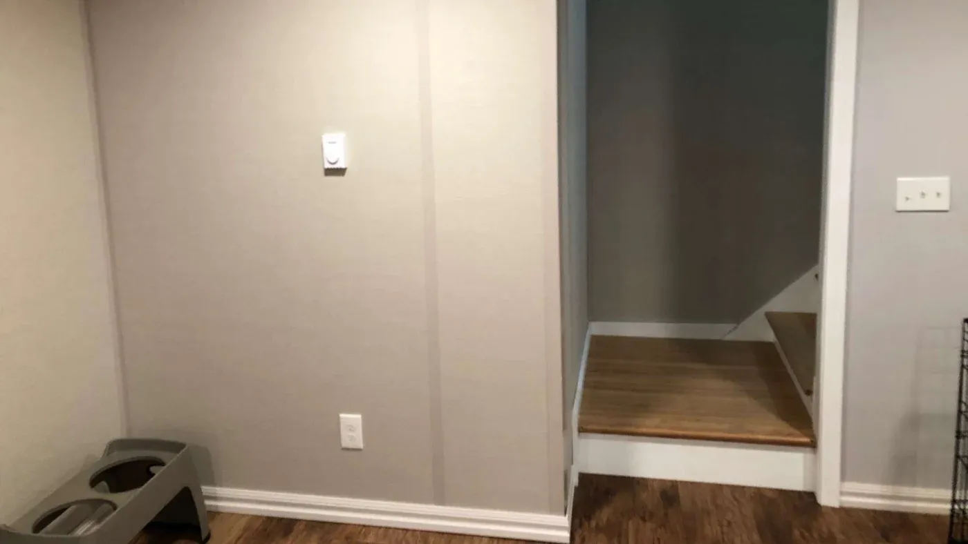 A cozy corner of a beige room with a dog bowl stand on the wooden floor. A thermostat and electrical outlet are on the wall. A small staircase leads up.