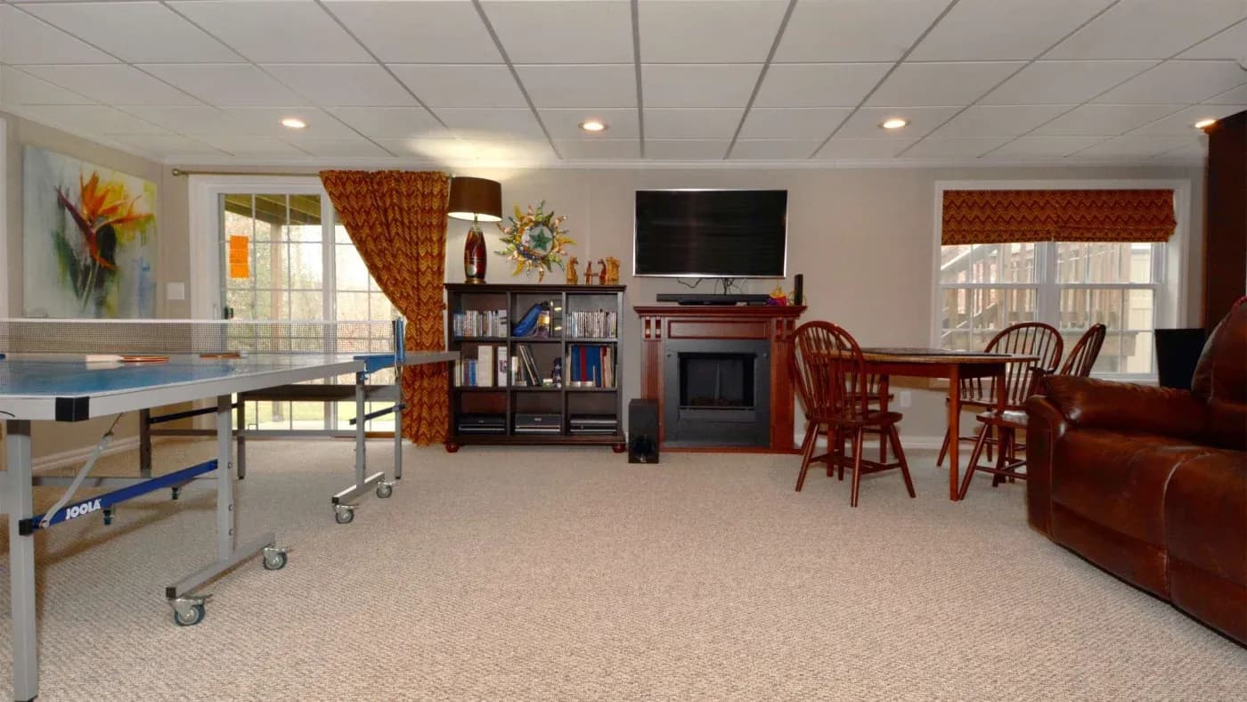 A cozy basement game room with a ping pong table, round wooden dining table, and leather couch. Shelves, wall art, and a TV above a fireplace add warmth.
