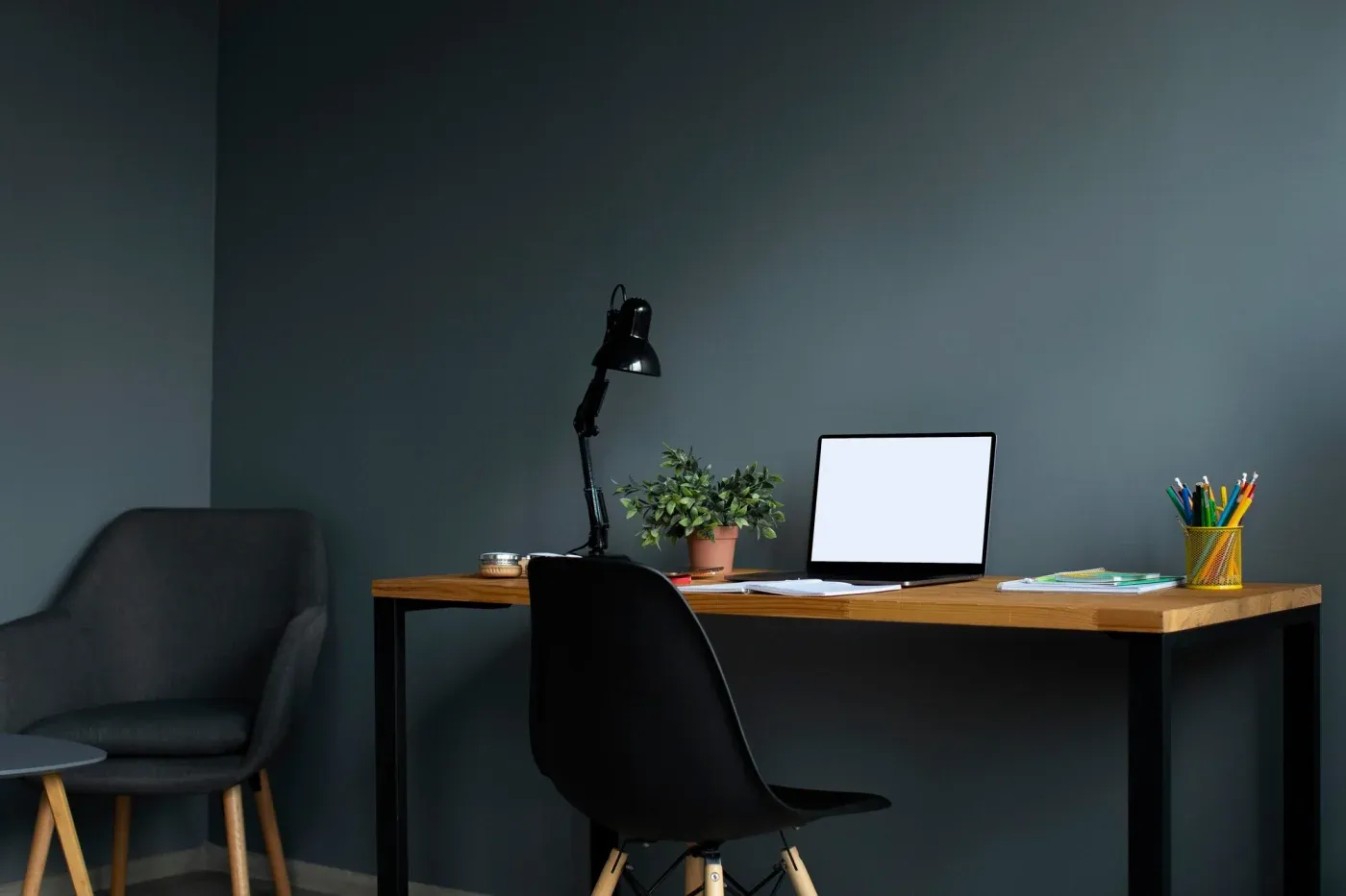 A minimalistic workspace with a wooden desk, black chair, laptop, desk lamp, potted plant, and colorful pens, set against a dark gray wall. Cozy and organized.