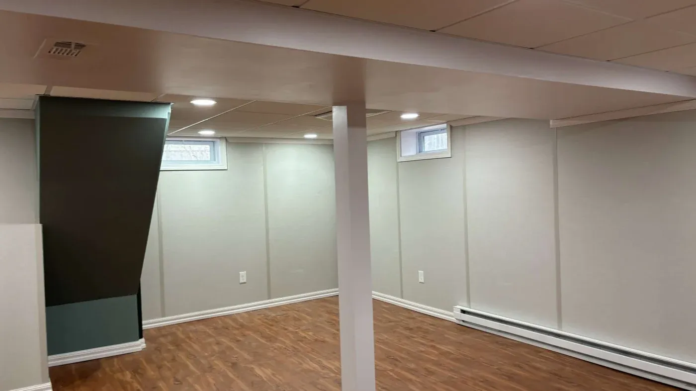 A finished basement with light gray walls, wooden flooring, and recessed lighting. Small windows provide natural light, creating a calm and open atmosphere.