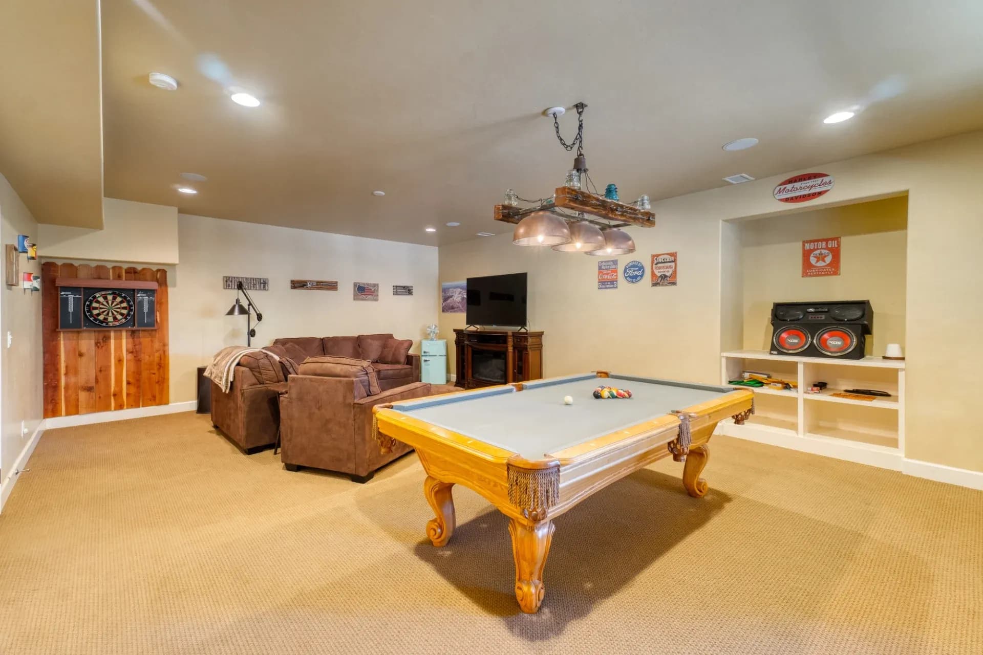 A cozy basement game room featuring a pool table under hanging lights, a plush brown sectional sofa, dartboard, retro stereo, and vintage decor on beige walls.