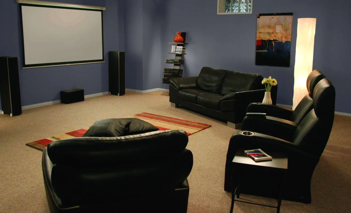 Cozy home theater with black leather seating, a large projector screen, and modern decor on a soft carpet. Warm lighting creates a relaxed ambiance.