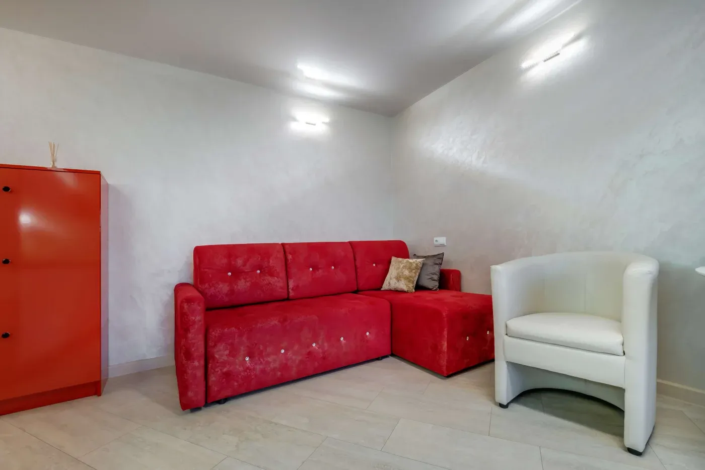 A modern room with a red sectional sofa and decorative pillows, adjacent to a white armchair and an orange cabinet. The walls are light gray.