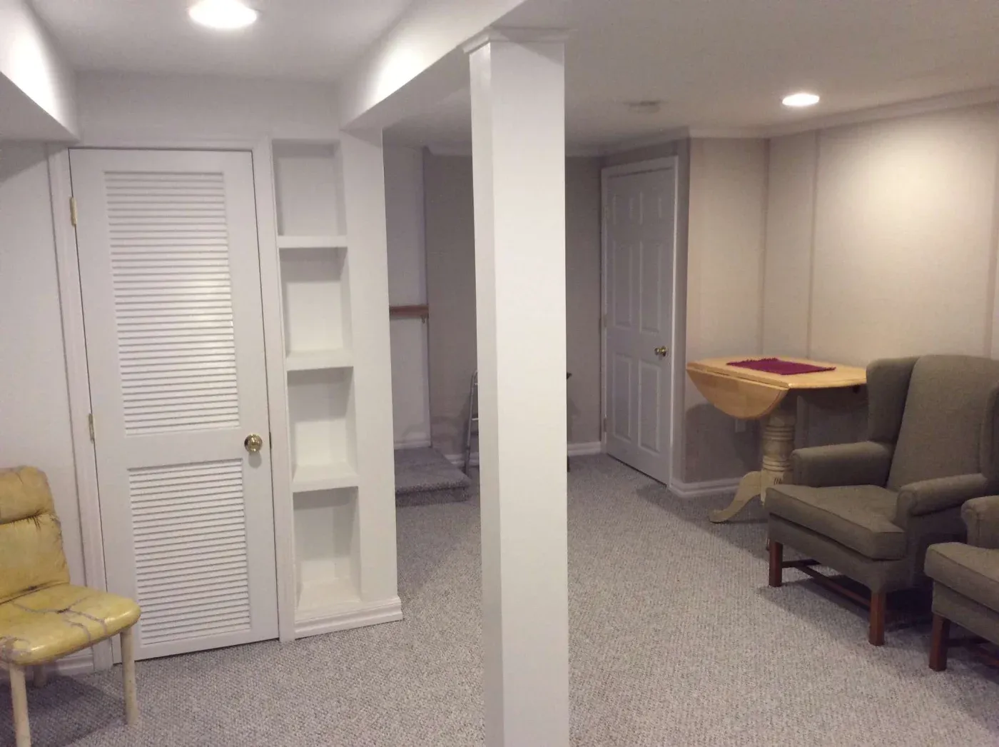 A cozy basement with white walls, carpeted floor, and recessed lighting. Features two armchairs, a drop-leaf table, a closet, and built-in shelves.