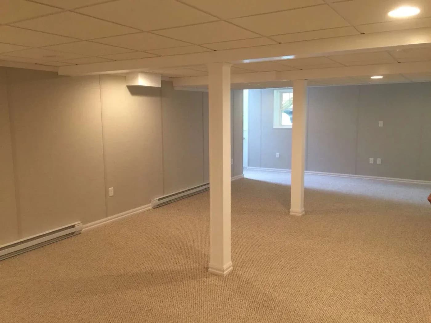 Spacious basement room with beige carpet, white support columns, recessed ceiling lights, and a small window with natural light entering from an exterior wall