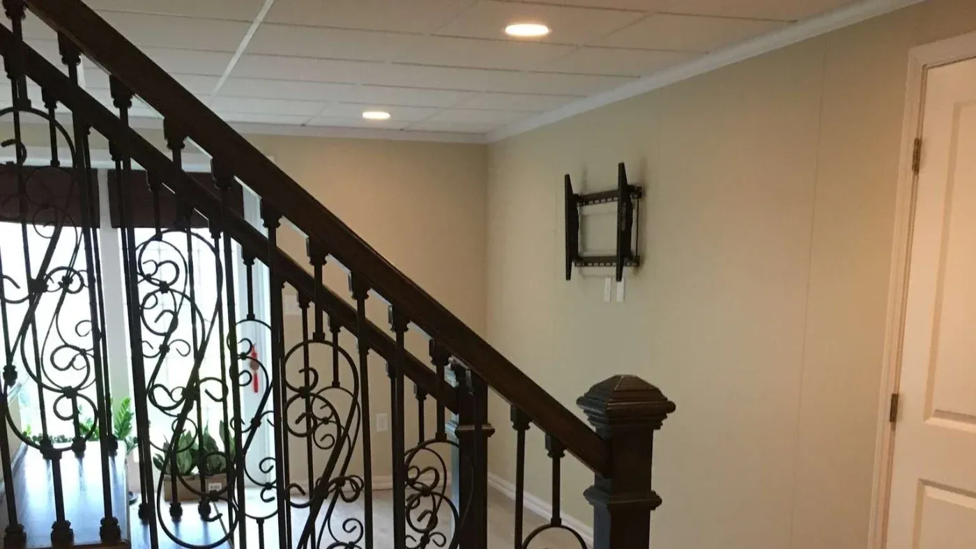 A beige hallway with a dark ornate iron staircase. Sunlight streams through a window with potted plants. Walls are plain, featuring an empty wall-mounted stand.