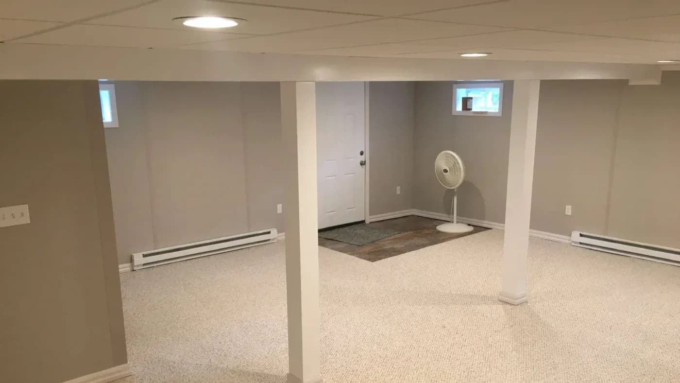 A spacious, empty basement with light beige carpet, gray walls, and white ceiling tiles. A white fan stands by a closed door, conveying a calm, minimalist tone.