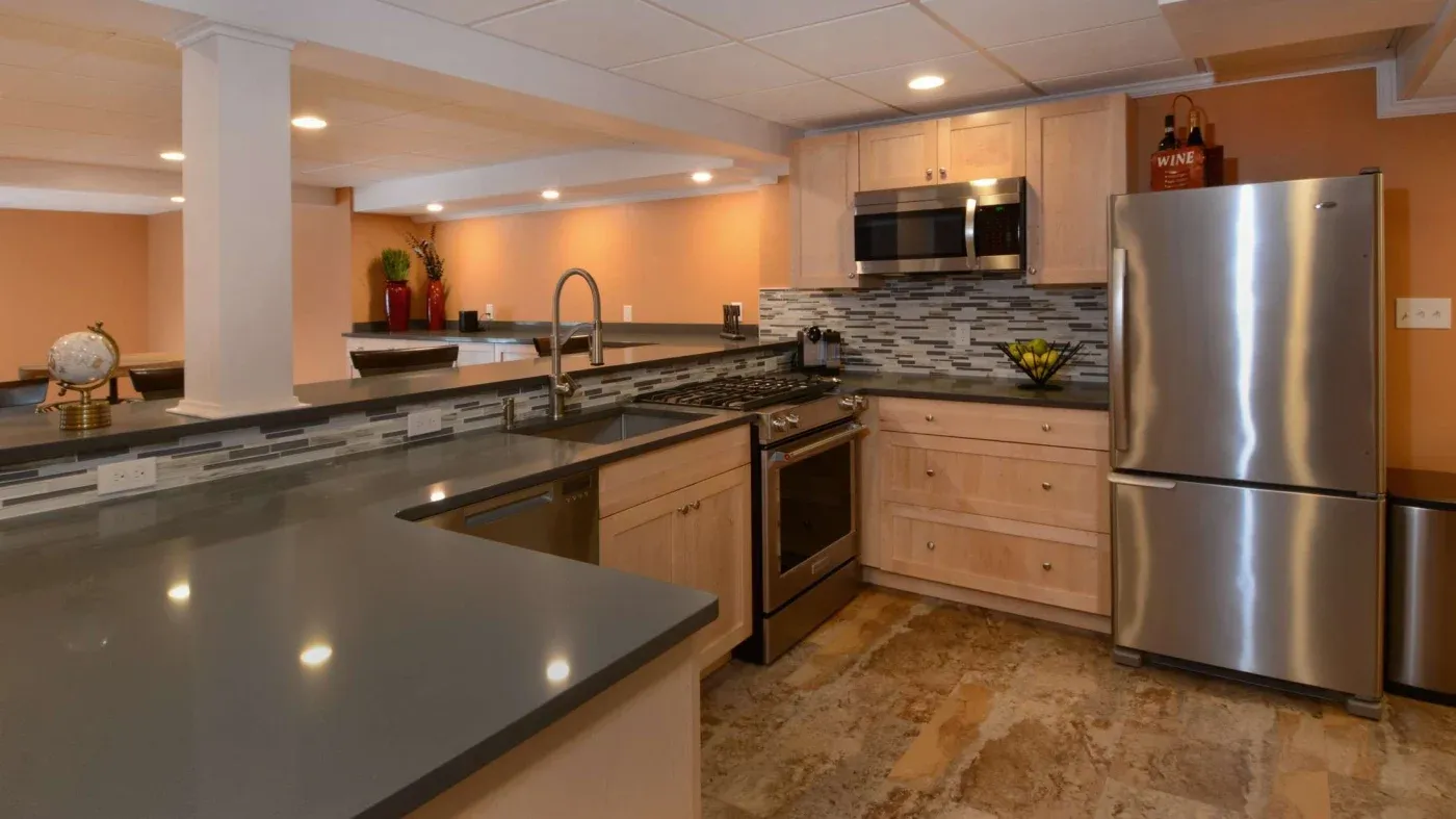 Modern kitchen with light wood cabinets, stainless steel appliances, and gray countertops. Warm lighting highlights the tile backsplash and earthy tones.