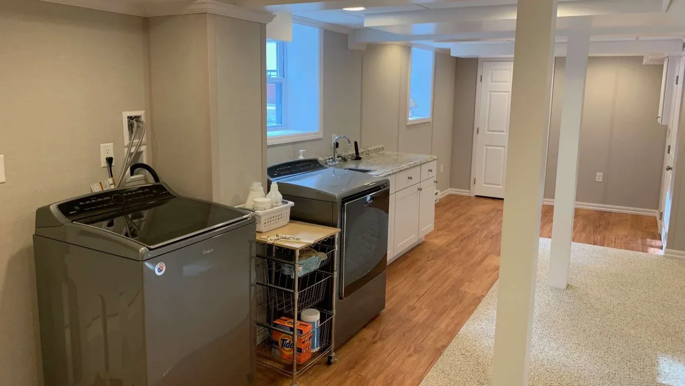 A well-lit basement laundry room featuring a washer and dryer, wooden floor, and white cabinetry. A metal rack holds laundry supplies, creating an organized space.