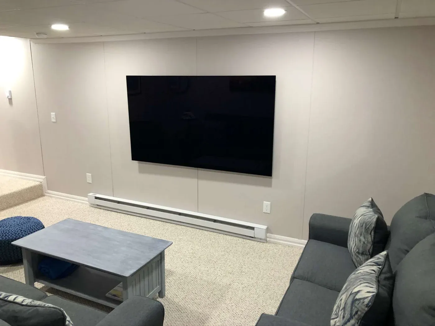 A cozy living room with a large black flat-screen TV mounted on a light gray wall. A gray couch with patterned pillows faces the TV, and a small blue-gray coffee table is in front, on a carpeted floor, creating a relaxed setting.