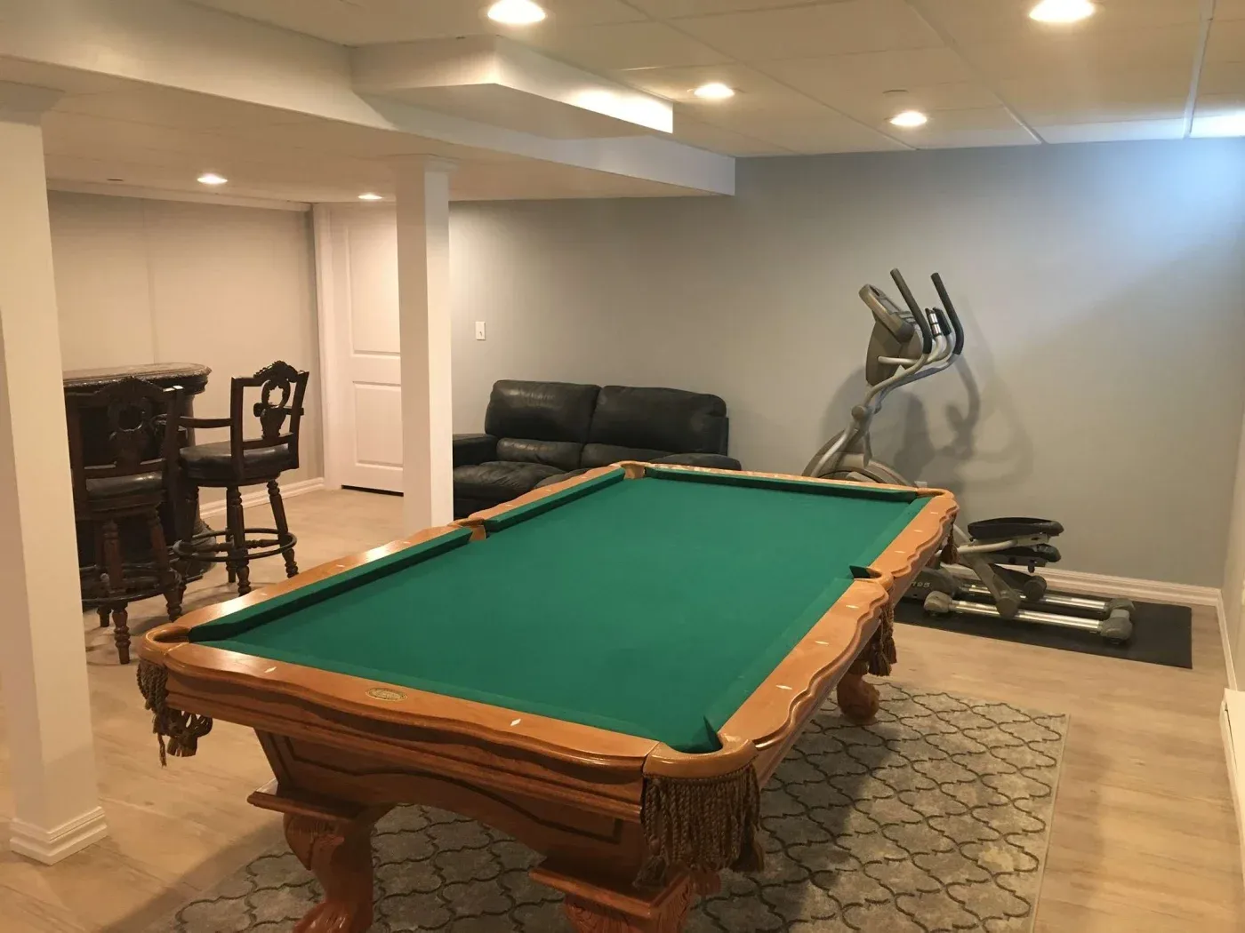A cozy basement features a billiards table with a green felt surface, a black leather couch against a pale wall, an exercise machine, and a small bar with stools.