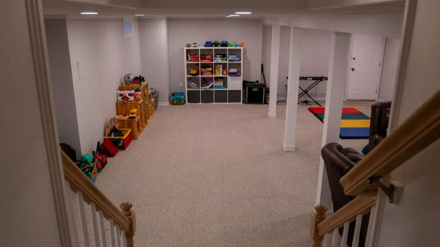 Spacious basement playroom with carpeted floor, shelves of toys, and a toy piano. Bright and organized, conveying a fun and welcoming atmosphere.