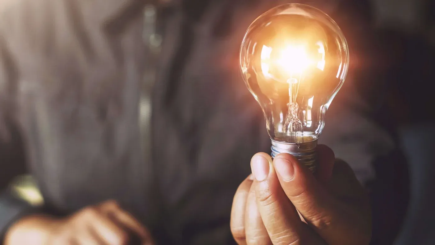 A person holds a lit light bulb in their hand, symbolizing innovation and ideas. The background is blurred, emphasizing the bright, glowing bulb.