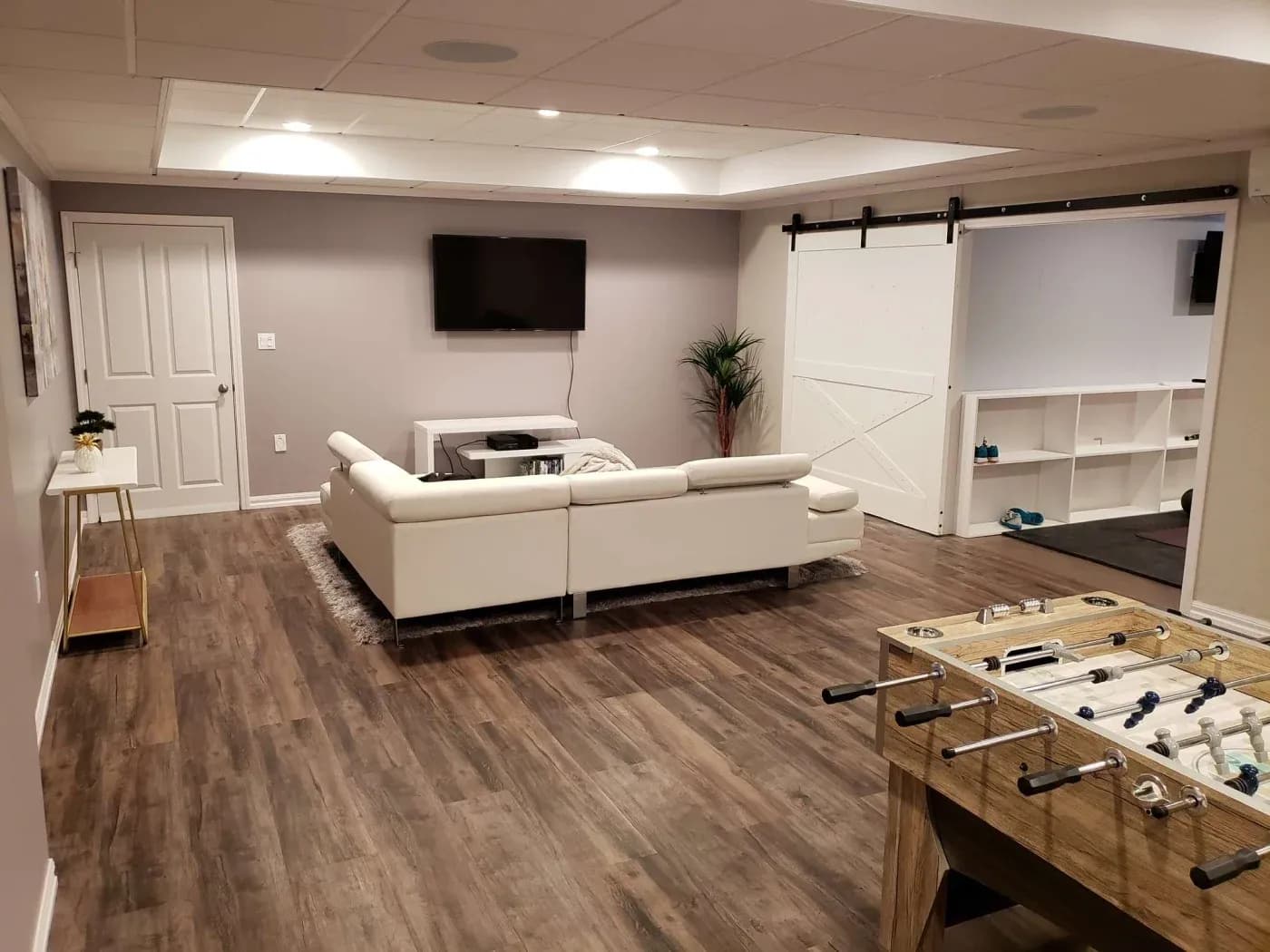 A modern basement with wood flooring features a cozy white sectional sofa facing a wall-mounted TV. A foosball table is in the foreground, and a sliding barn door reveals a gym space. A potted plant adds a touch of greenery.