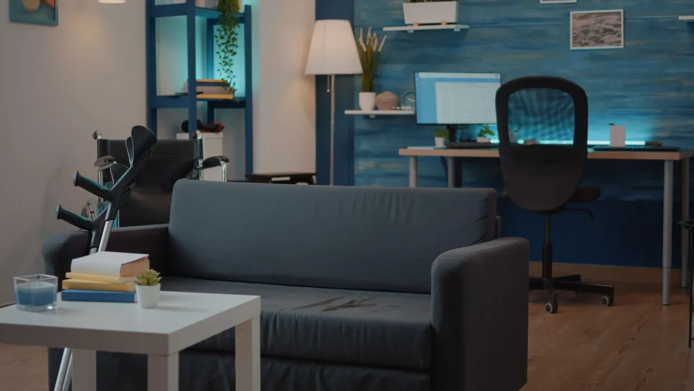 Cozy living room with a gray sofa, white table, crutches, and a wheelchair. Blue walls, a lit lamp, shelves, and a home office in the background.