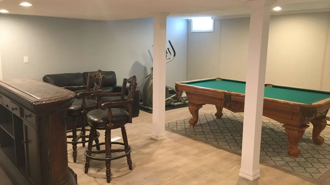 A cozy basement recreation area with light wood floors features a billiards table on a patterned rug, bar with ornate stools, sofa, and exercise bike.