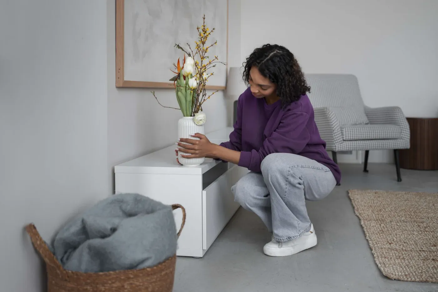 A person in a purple sweater and jeans arranges flowers in a white vase on a white cabinet. The room is calm, with a gray chair and woven basket.